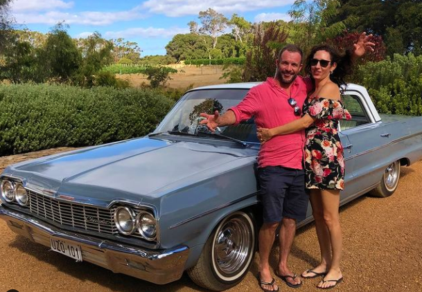 A couple poses happily in front of a classic 1964 Chevy car amidst a scenic vineyard landscape in the Margaret River Wine Region.