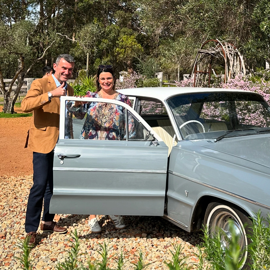 A man and a woman standing next to a classic 1964 Chevrolet during a Belair Affair wine tour.