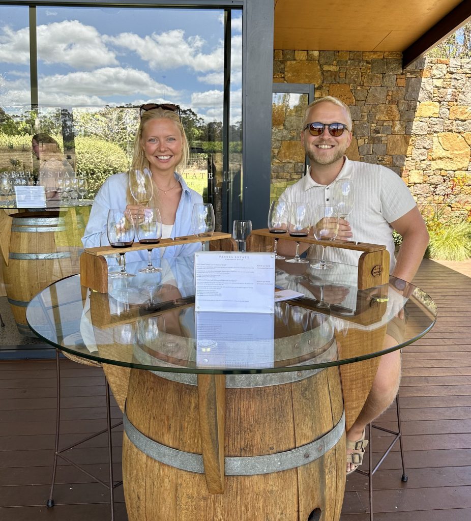 A woman and a man enjoying a wine tasting at a vineyard, seated at a glass table with wine glasses and wooden barrels in the background.